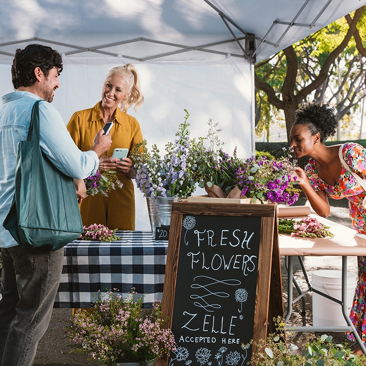 People buying flowers at an outdoor booth with a Zelle accepted here sign.
