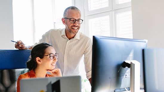 A smiling man with glasses and a white shirt leans over a woman with glasses and an orange top, as they look at a computer monitor together in a bright office.