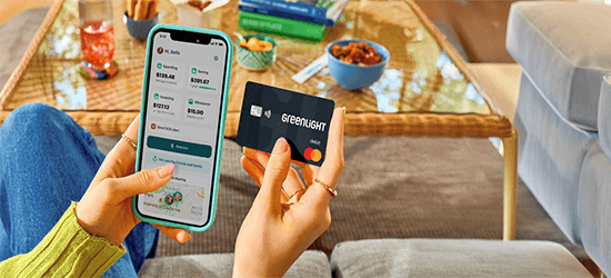 A person sitting indoors holds a smartphone in one hand displaying the Greenlight app with spending, saving, and investing balances, and a black Greenlight debit card in the other. A coffee table with snacks, books, and a drink is visible in the background, creating a casual home setting.
