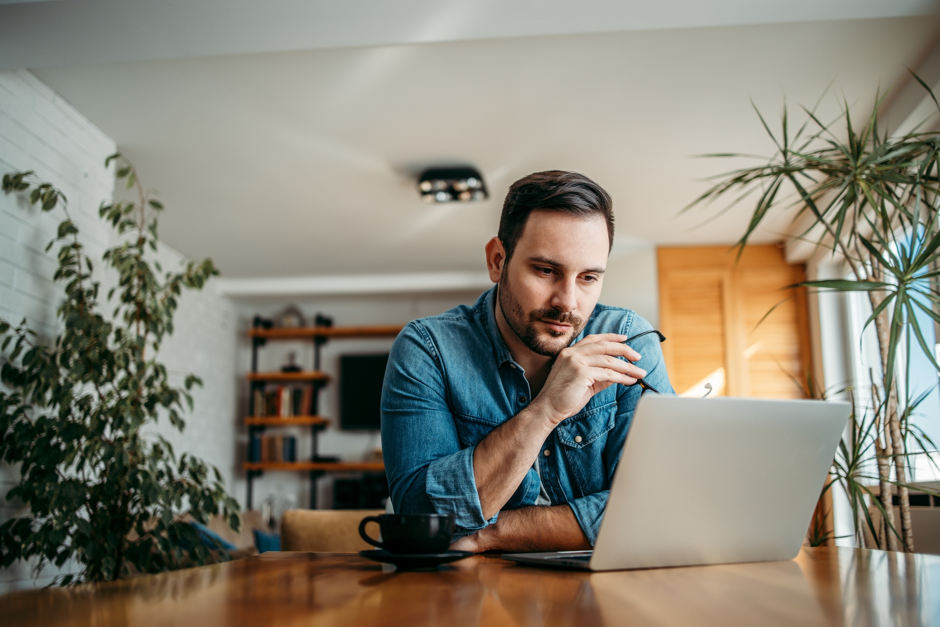 A man in a denim shirt sits at a wooden table, looking thoughtfully at a laptop while holding a pair of glasses.