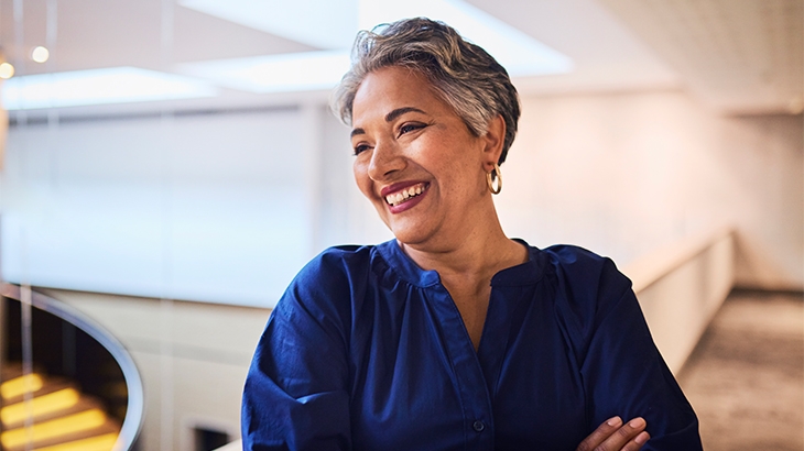 A smiling woman with short gray hair and a dark blue shirt stands with her arms crossed in a bright, modern indoor office space.