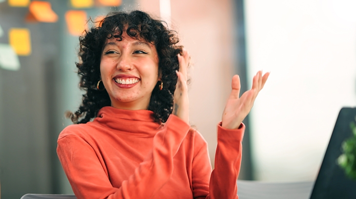 A happy woman with curly hair and a coral turtleneck is clapping in an office setting.
