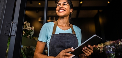 Smiling small business owner in an apron holds a tablet while standing in the doorway of her shop.