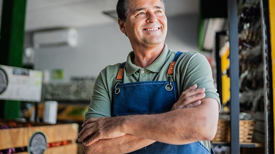 A smiling small business owner wearing an apron stands with arms crossed in his shop.
