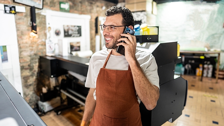 A smiling man with glasses and a brown apron talks on a cell phone inside a print shop, standing next to a large-format printer.