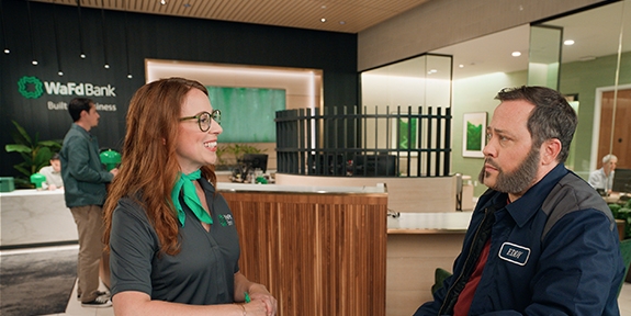 A smiling WaFd Bank employee wearing a green scarf talks to a male customer in a work jacket inside a WaFd branch.