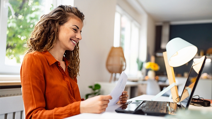 A smiling woman with long, curly brown hair is sitting at a desk indoors, looking down at a document in her hands with a laptop and desk lamp in front of her.