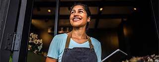 Smiling small business owner in an apron holds a tablet while standing in the doorway of her shop.