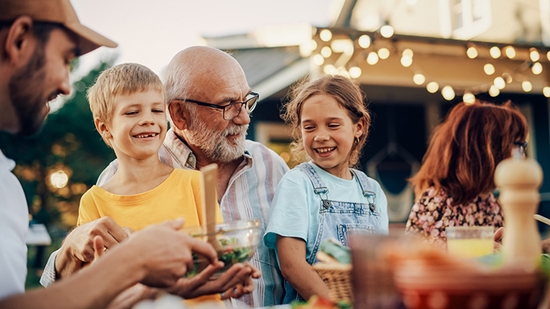 A multi-generational family smiles and talks together while sharing a meal at an outdoor patio table decorated with string lights.