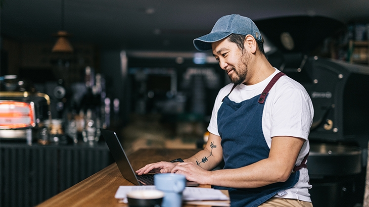 A business owner wearing an apron and a baseball cap, working on a laptop.
