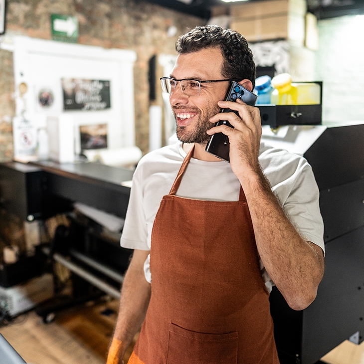 A smiling man with glasses and a brown apron talks on a cell phone inside a print shop, standing next to a large-format printer.