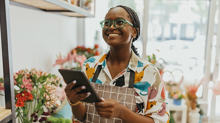 A cheerful Black woman with braided hair, wearing glasses and a patterned shirt under an apron, stands in a flower shop, holding a tablet and smiling.