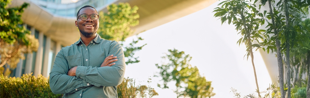 A smiling man with glasses and arms crossed stands outdoors in front of a modern glass building and a skybridge.