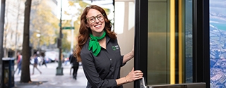 A smiling woman in a gray WaFd Bank uniform and green scarf holds open a glass door to a bank branch.