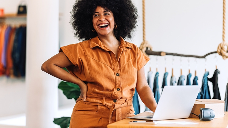 Woman laughing in clothing shop with laptop on counter.