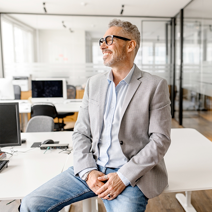 A smiling, gray-haired businessman in a light gray blazer and jeans sits on a desk in a modern office, looking off to the side.