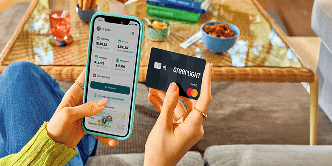 A person sitting indoors holds a smartphone in one hand displaying the Greenlight app with spending, saving, and investing balances, and a black Greenlight debit card in the other. A coffee table with snacks, books, and a drink is visible in the background, creating a casual home setting.