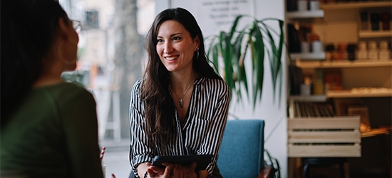 A woman in a striped shirt smiles while holding a tablet and talking to a colleague in a bright, modern cafe setting.