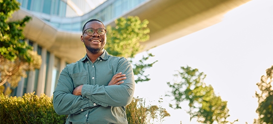 A smiling man with glasses and arms crossed stands outdoors in front of a modern glass building and a skybridge.