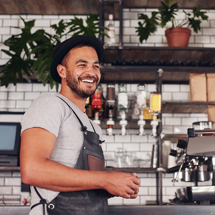 A smiling man in a black apron and fedora standing behind a counter with an espresso machine in a cafe.