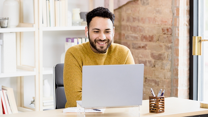 A smiling man with a beard, wearing a mustard yellow sweater, sits at a desk in a modern office looking at a laptop.