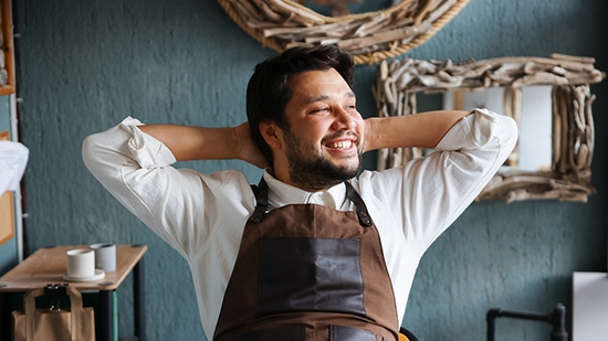 A smiling man in a brown artisan apron leans back with his hands behind his head, looking relaxed in his workshop.