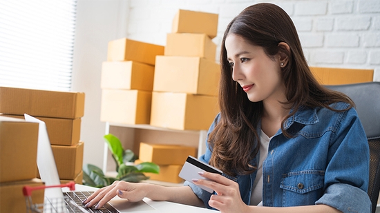 A woman in a denim shirt sits at a laptop holding a credit card, surrounded by stacks of shipping boxes.