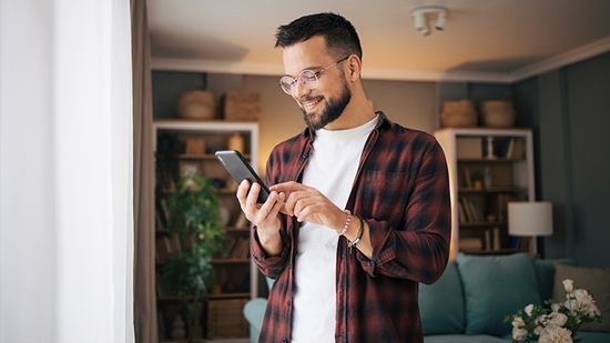 Un hombre con barba y gafas mira su teléfono inteligente y sonríe mientras está junto a una ventana.