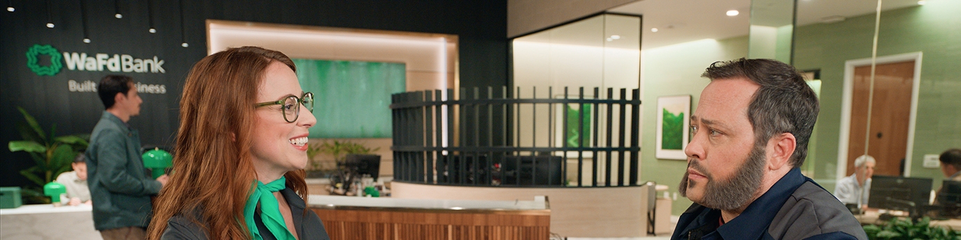 A smiling WaFd Bank employee wearing a green scarf talks to a male customer in a work jacket inside a WaFd branch.