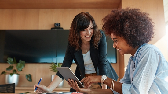 Two businesswomen smiling while looking at a digital tablet and taking notes in a modern meeting room.