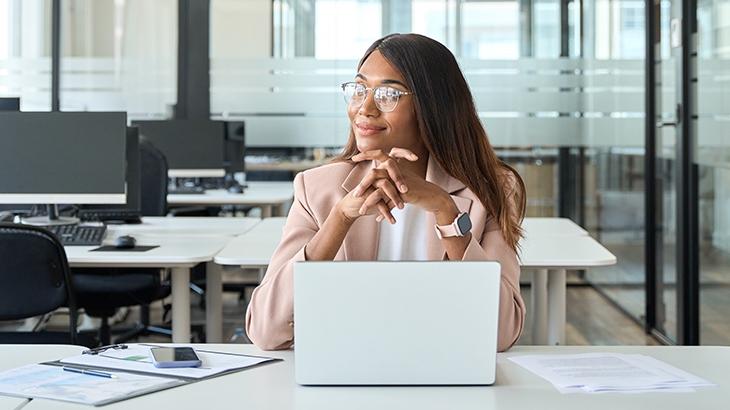 Smiling woman wearing glasses, sitting at an office desk with a laptop and papers.
