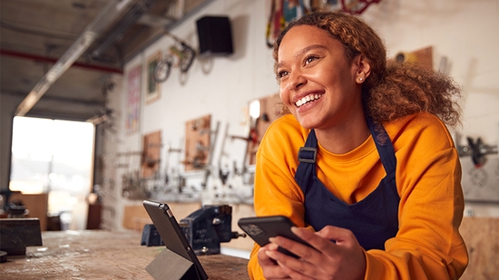 A smiling young woman in an apron and a yellow sweater holds a smartphone while leaning over a workbench with a tablet in a workshop.