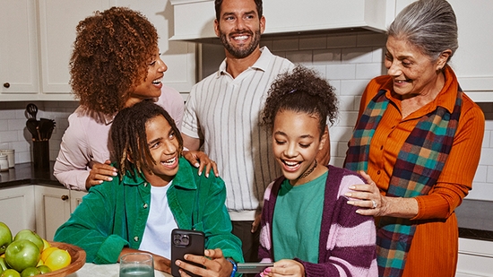 A multi-generational family smiles together in a kitchen while two teenagers look at a smartphone and a Greenlight debit card.
