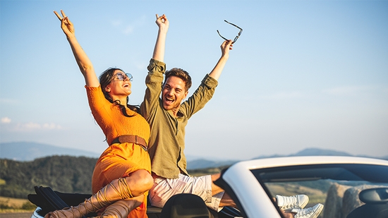 Man and woman sitting on a car celebrating with hands in the air.