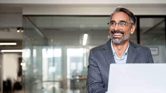 Smiling businessman with gray hair and glasses working on a laptop in a modern office.