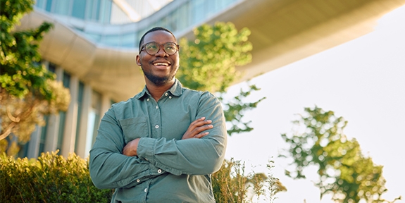 A smiling man with glasses and arms crossed stands outdoors in front of a modern glass building and a skybridge.