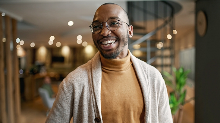 A smiling Black man wearing glasses, a tan turtleneck, and a light-colored cardigan stands in a modern, well-lit office.