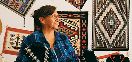 An older woman in a plaid shirt stands in a shop featuring Native American jewelry and woven rugs on the wall.