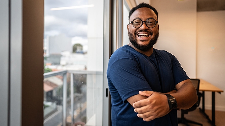 Happy man in a blue shirt and glasses, standing by a window with his arms crossed in an office.